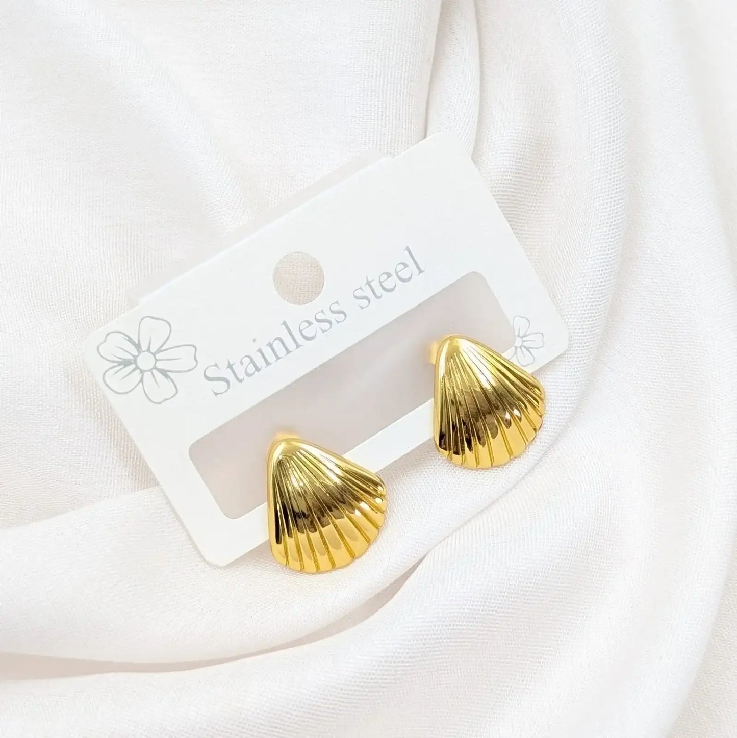 Gold shell-shaped earrings on a white card with 'Stainless steel' text, against a white fabric background.