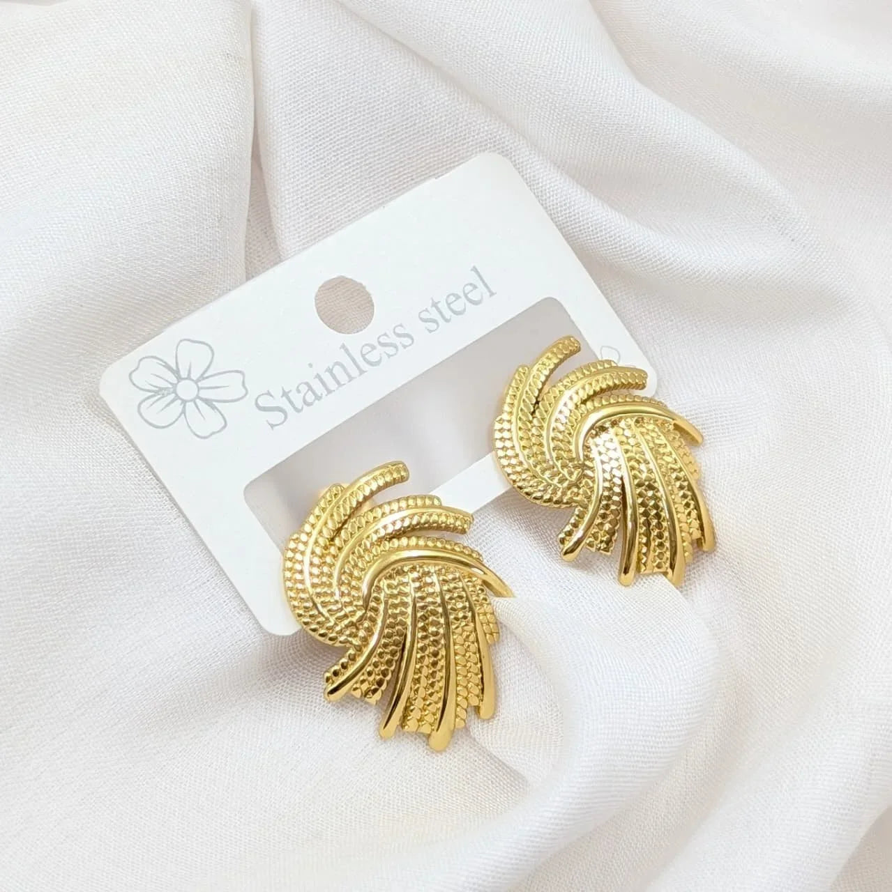 Gold earrings on a white card with 'Stainless steel' text, placed on a white fabric background.