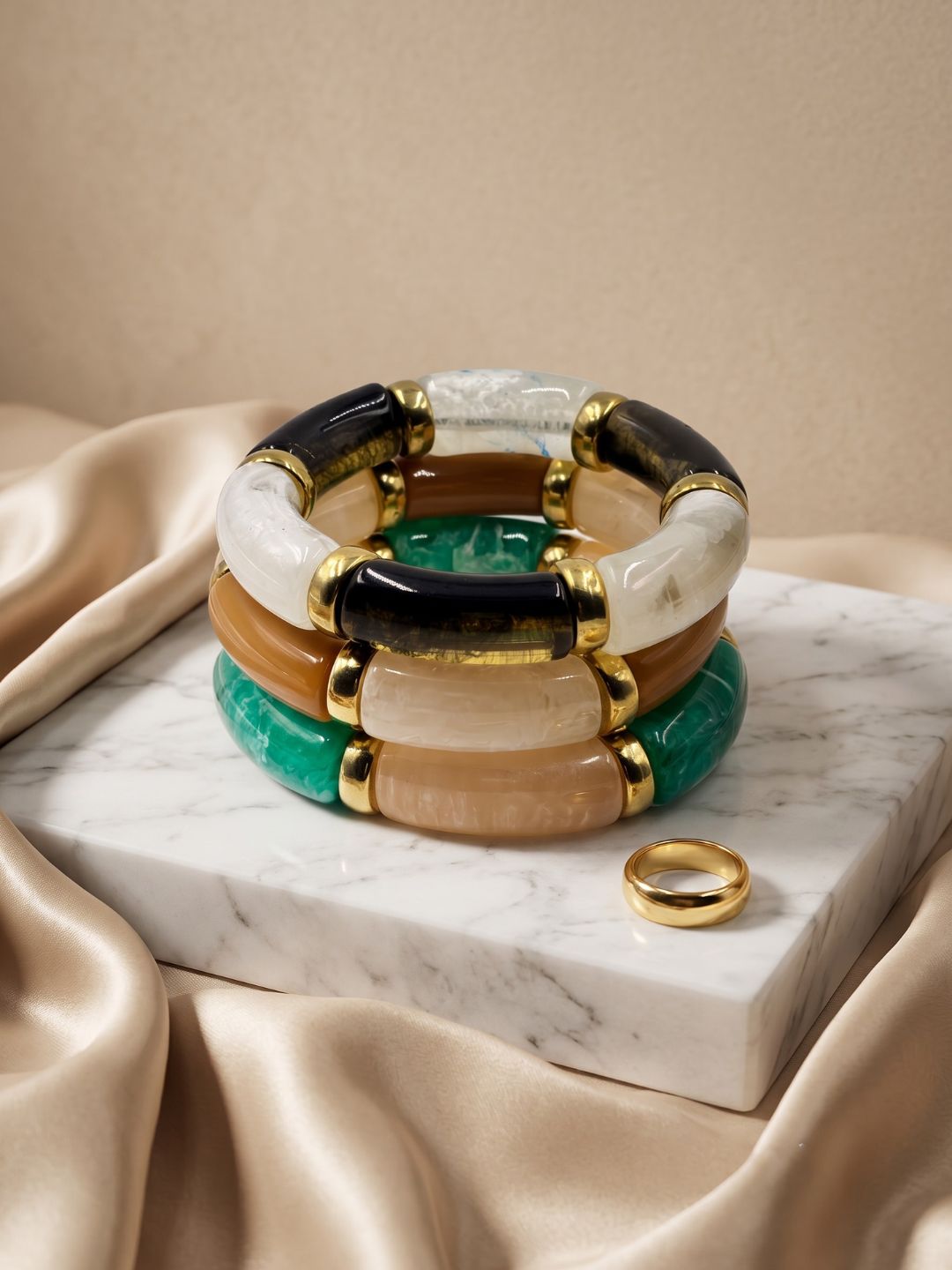 A stack of chunky bangle bracelets with alternating black, white, green, and tan stone sections, accented with gold-toned metal caps, displayed on a white marble block with draped champagne satin fabric.