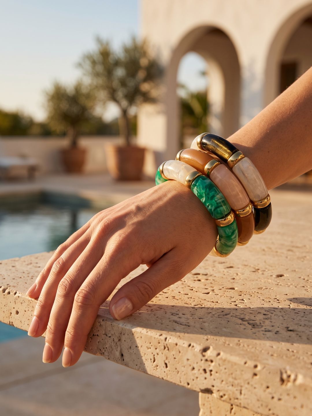 A close-up of a person's hand resting on a stone ledge by a pool, wearing a stack of chunky bangles. The bracelets include green marbled stone, cream-colored stone, tan stone, and polished gold-toned metal.