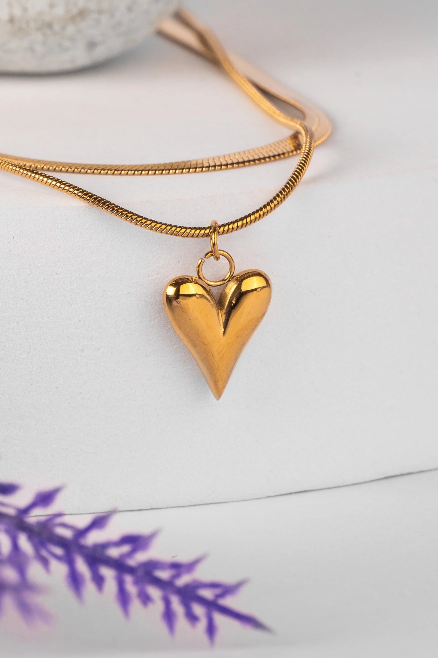 Gold heart-shaped pendant on a gold chain against a white background with purple leaves.