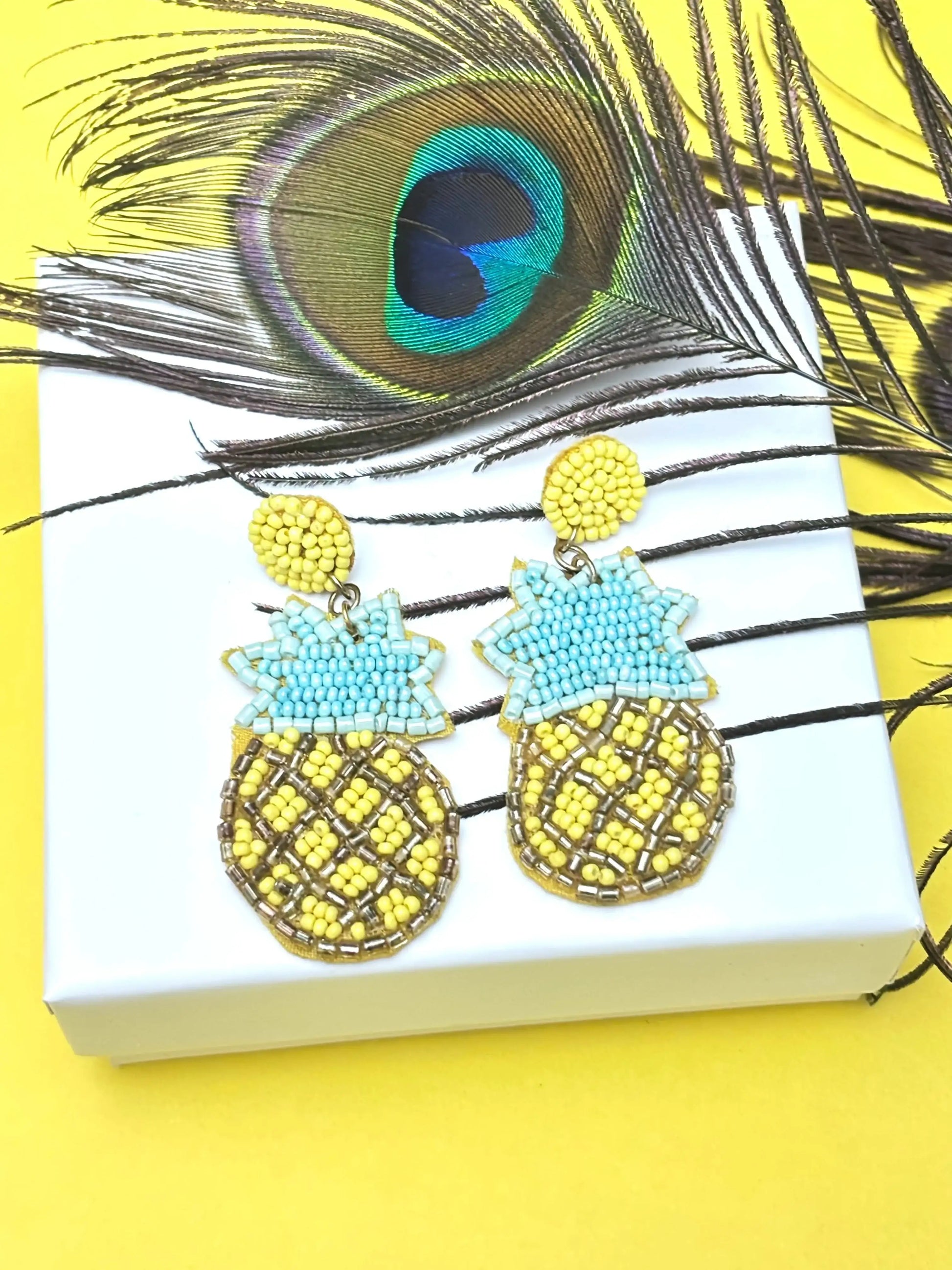 Pineapple-shaped earrings on a white box with peacock feathers in the background