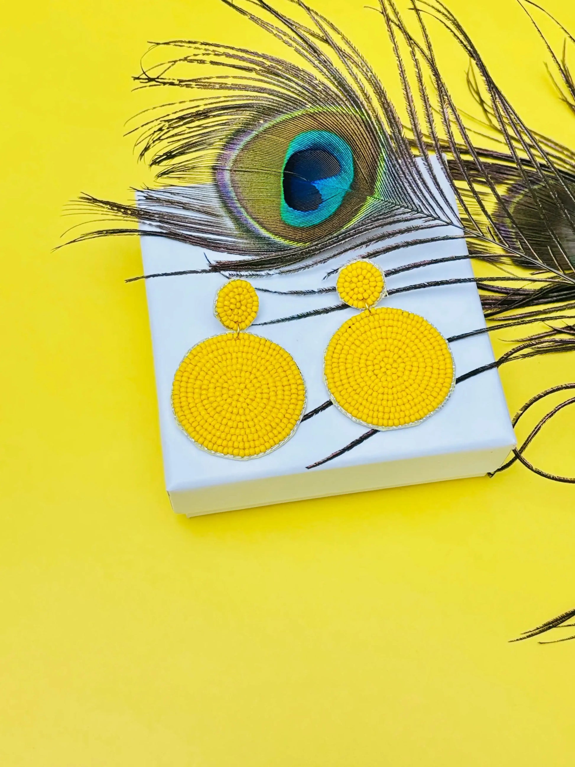 Yellow earrings on a white box with a peacock feather on a yellow background