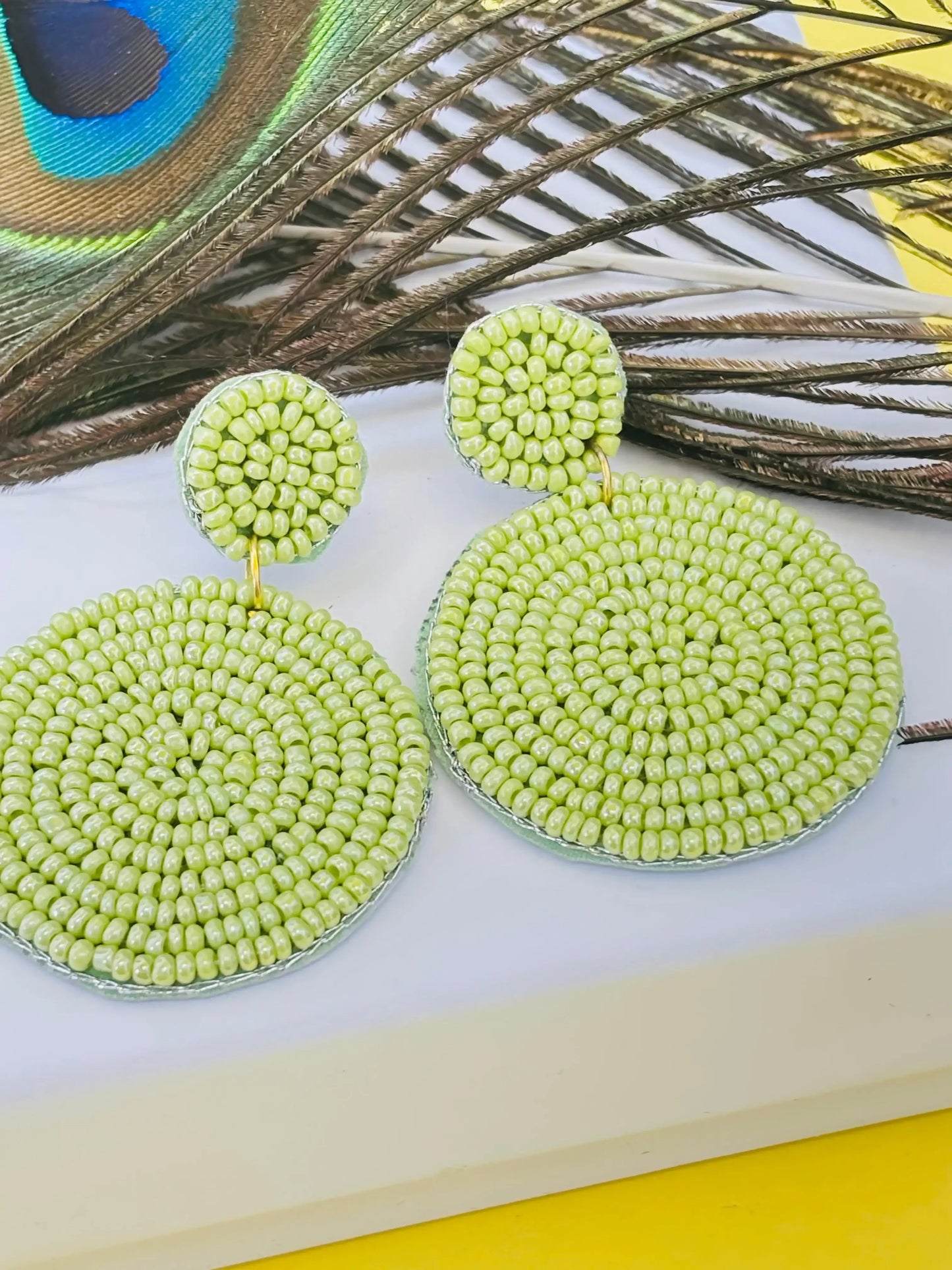 Green beaded earrings on a white surface with peacock feathers in the background