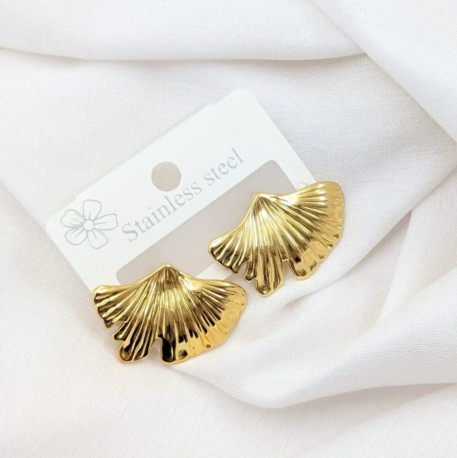 Gold shell-shaped earrings on a white card with 'Stainless steel' text, placed on a white fabric background.