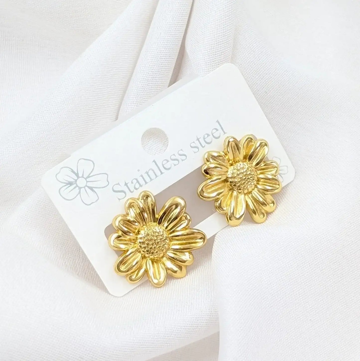 Gold flower-shaped earrings on a white card with 'Stainless steel' text, against a white fabric background.