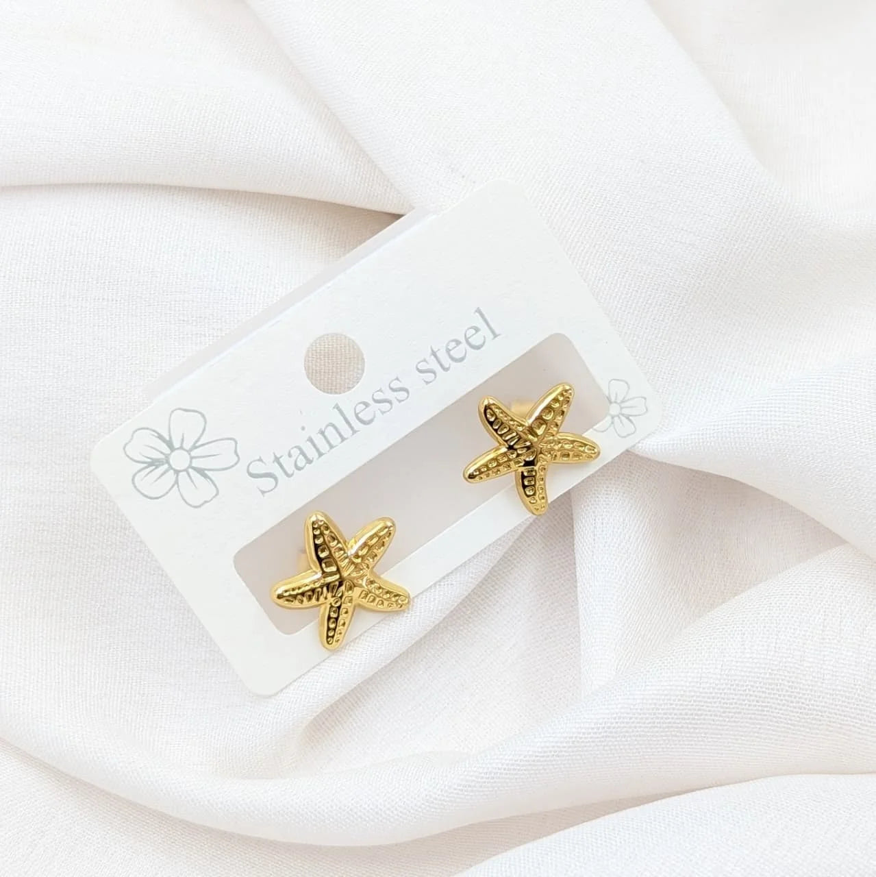 Gold starfish earrings on a white card with 'Stainless steel' text, against a white fabric background.