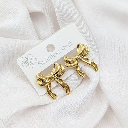 Gold bow-shaped earrings on a white card with 'Stainless steel' text, placed on a white fabric background.